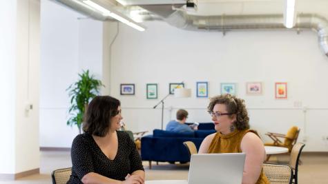 Two people sit at a table in front of a laptop.