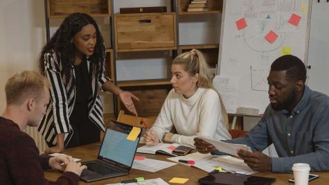 A group of four people at a table having a conversation. Papers and laptops cover the table.