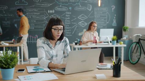 A classroom with a chalk board in the background. In the foreground, People work at laptops at desks. 