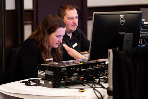 Two people sit at a tech desk, turning dials on a soundboard and looking into monitors