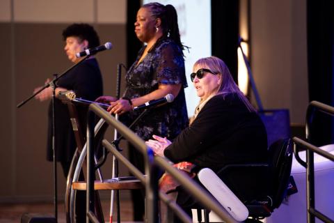 Several speakers stand on a stage. One of them is at a podium, another is in a mobility aid in front of a microphone, and the other is signing for the crowd