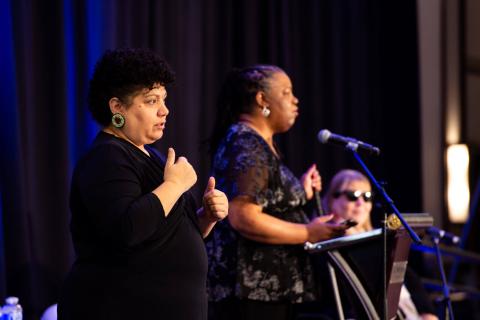 Three speakers stand onstage. In the foreground, the interpreter signs to the audience