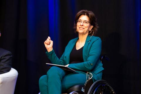 A person in a wheelchair speaks to the audience with a clipboard in their lap