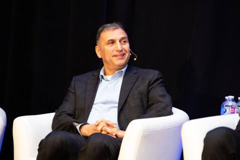 A speaker sits onstage in a white chair smiling at another speaker