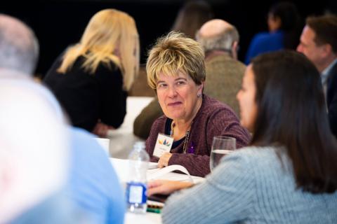 The audience sits at tables. In the foreground, two people speak to each other. Water bottles and glasses accompany them at the tables
