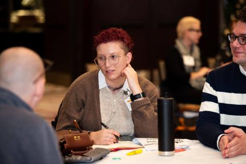 A group of three sit at a table around papers and drinks, talking and smiling to one another