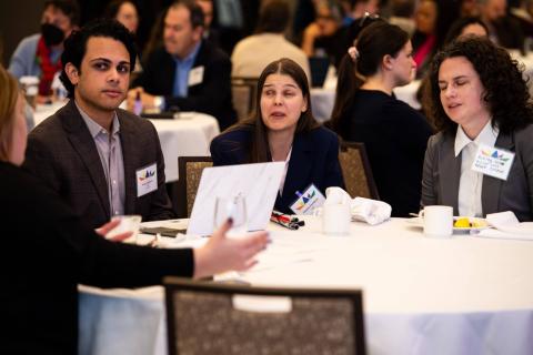 Four people sit around a table with a white tablecloth. Mugs of coffee and tea, plates of food, and paperwork are scattered along its surface as the four people talk with one another