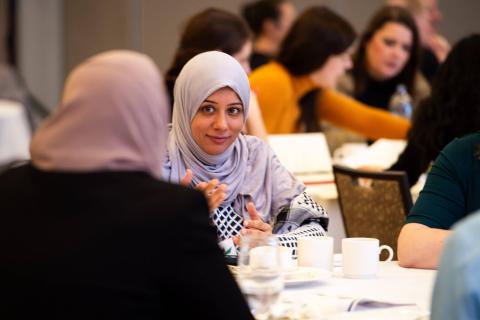 Two people talk at a table. One of them faces the camera while the other has their back to it