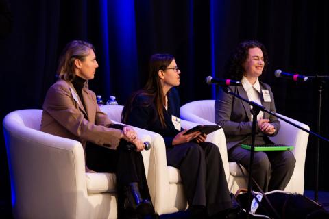 Three speakers sit in white chairs onstage. Microphones on stands are facing them. One of the people speaks to the audience while the other two watch them
