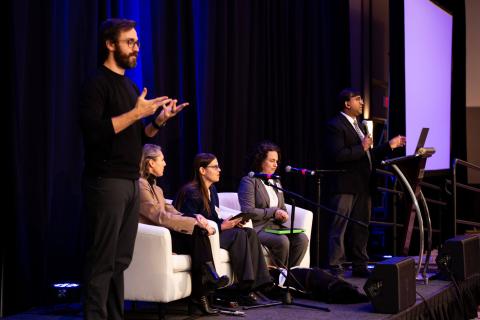 Three speakers sit on white chairs onstage while another stands and speaks into a microphone at a podium. A sign language interpreter stands in the foreground of the photo, signing to the audience