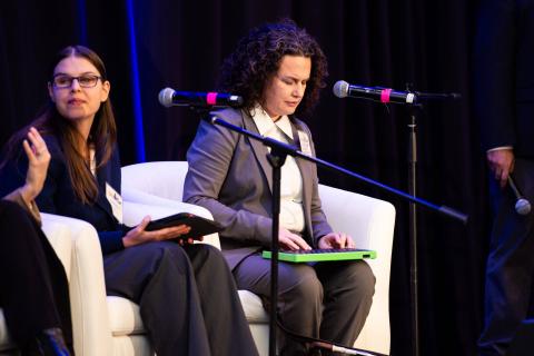 Three speakers sit onstage in white chairs. One of the speakers types on a tablet