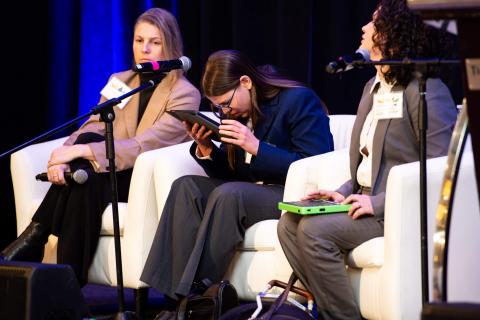 Three speakers sit in white chairs onstage. The middle figure holds a tablet up to look closely while another sits with their tablet in their lap