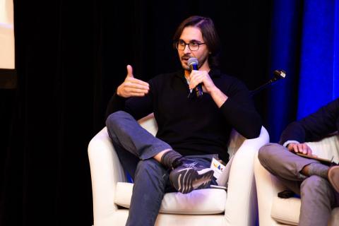 Two people sit in white chairs onstage. One speaker holds a microphone and speaks to the audience