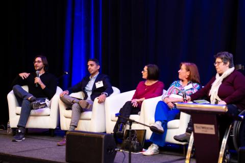 Five speakers sit onstage, four in white chairs and one in a wheelchair. Four of them watch one speak into a microphone