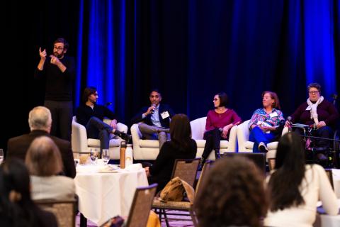 An audience perspective of the stage. Multiple audience members listen to a speaker onstage. Five speakers sit onstage while a sign language interpreter signs to the audience on the left hand side