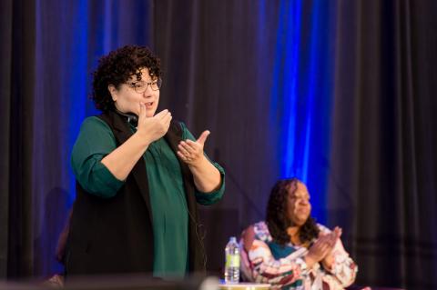 A speaker onstage in front of a blue curtain while an interpreter signs to the audience close by.
