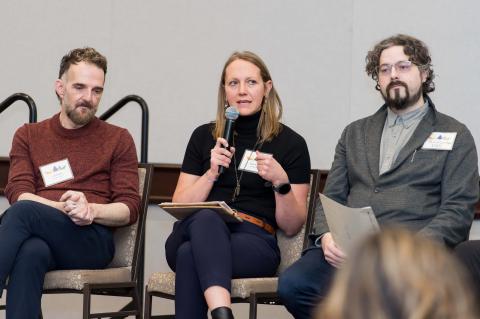 Three speakers wearing nametags sit in chairs on a stage. One of them speaks into a microphone. 