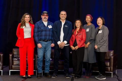 A group of six speakers with name tags stand on stage in front of a blue curtain 