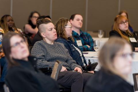 A crowd of people sit at white tables staring ahead at the stage