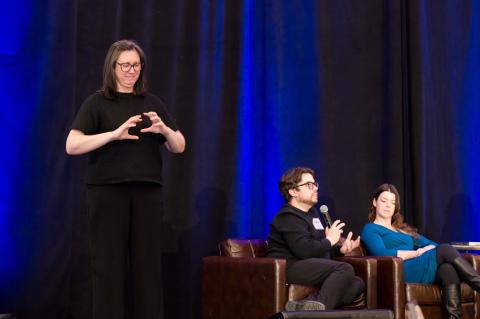Two speakers sit onstage in front of a blue curtain. An interpreter stands beside them signing to the audience.
