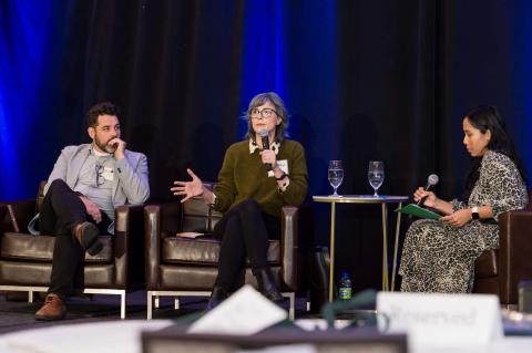 Three speakers sit on a stage in front of a blue curtain. Two of them face another, who speaks into a microphone.
