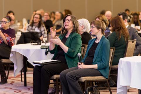 A crowd of people sitting at tables with white tablecloths. An audience member addresses the speakers on stage.