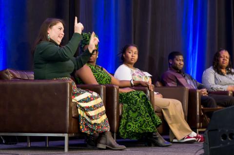 Five speakers sit onstage in brown chairs in front of a blue curtain. Four of them watch on as the fifth speaker speaks to the audience. 