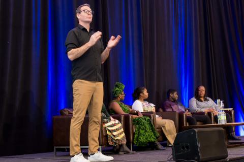 Five speakers sit onstage in front of a blue curtain. A sign language interpreter translates for the audience in the foreground.