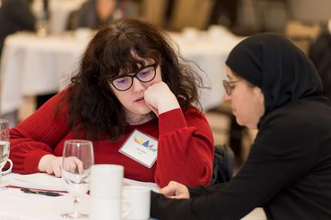 Two people at a table. They stare down at a phone and have a conversation.