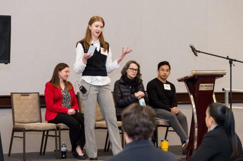 Three speakers sit on stage in a room with white walls. A fourth speaker stands and addresses the crowd.