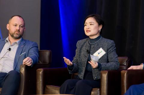 A speaker addresses the audience while sitting on stage. Another speaker sits to the left.
