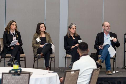 Four speakers sit in chairs on stage with name tags on.