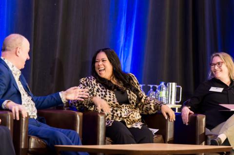 Three speakers sit on stage in front of a blue curtain. One speaker laughs at another's joke while the other watches on. 