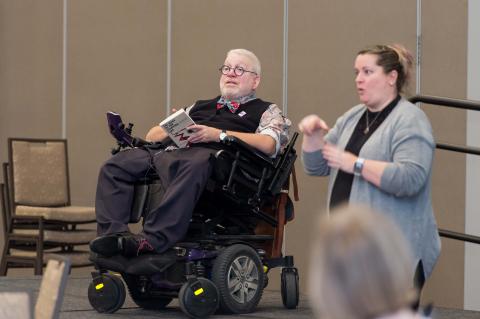 A person in a motorized wheelchair talks to the audience from a stage. An interpreter signs to the audience beside them.