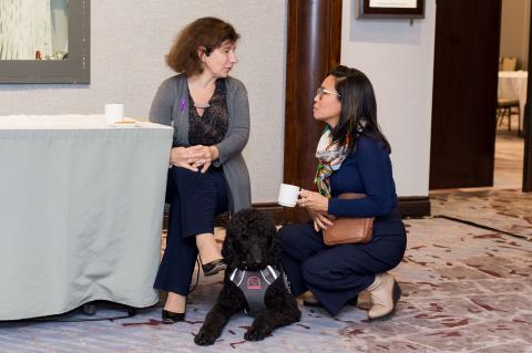 Two people talk with one another at a table with a white tablecloth. A service dog with black curly hair sits at their feet.