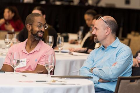 Two audience members sit at a table with water glasses. They smile and talk with each other. 