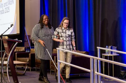 Two people walk off-stage. One uses a white cane. They smile as they leave.