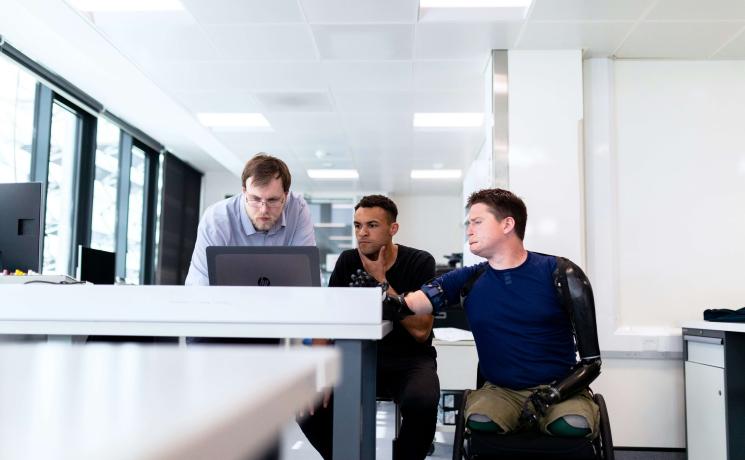 colleagues working at a computer; on is standing, one is seated in a chair and one is seated in a wheelchair 