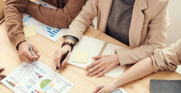 A group of people at a table. They are surrounding charts and hold pens to mark up their papers.