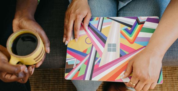 A laptop with a colourful Pride cover laying on someone's lap. A mug of coffee held in the hands of another.
