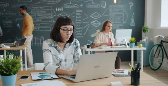 A classroom with a chalk board in the background. In the foreground, People work at laptops at desks. 