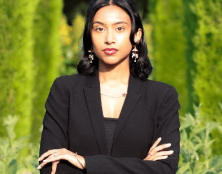 Portrait of Ronessa Dass standing outdoors against a green leafy background, wearing a black outfit with arms crossed and looking at the camera.